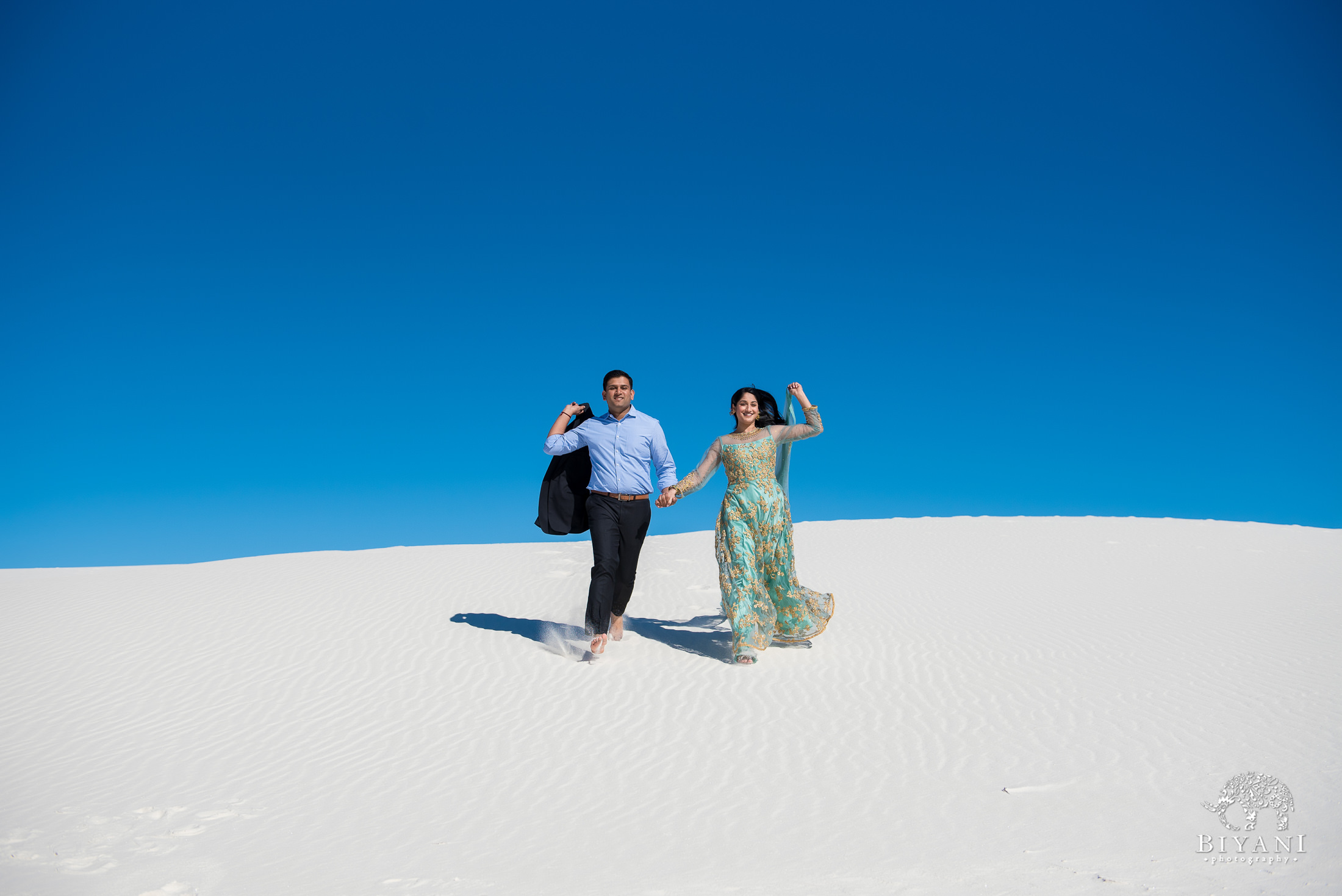 couple runs down clean white sand dune holding hands during a bright afternoon in New Mexico