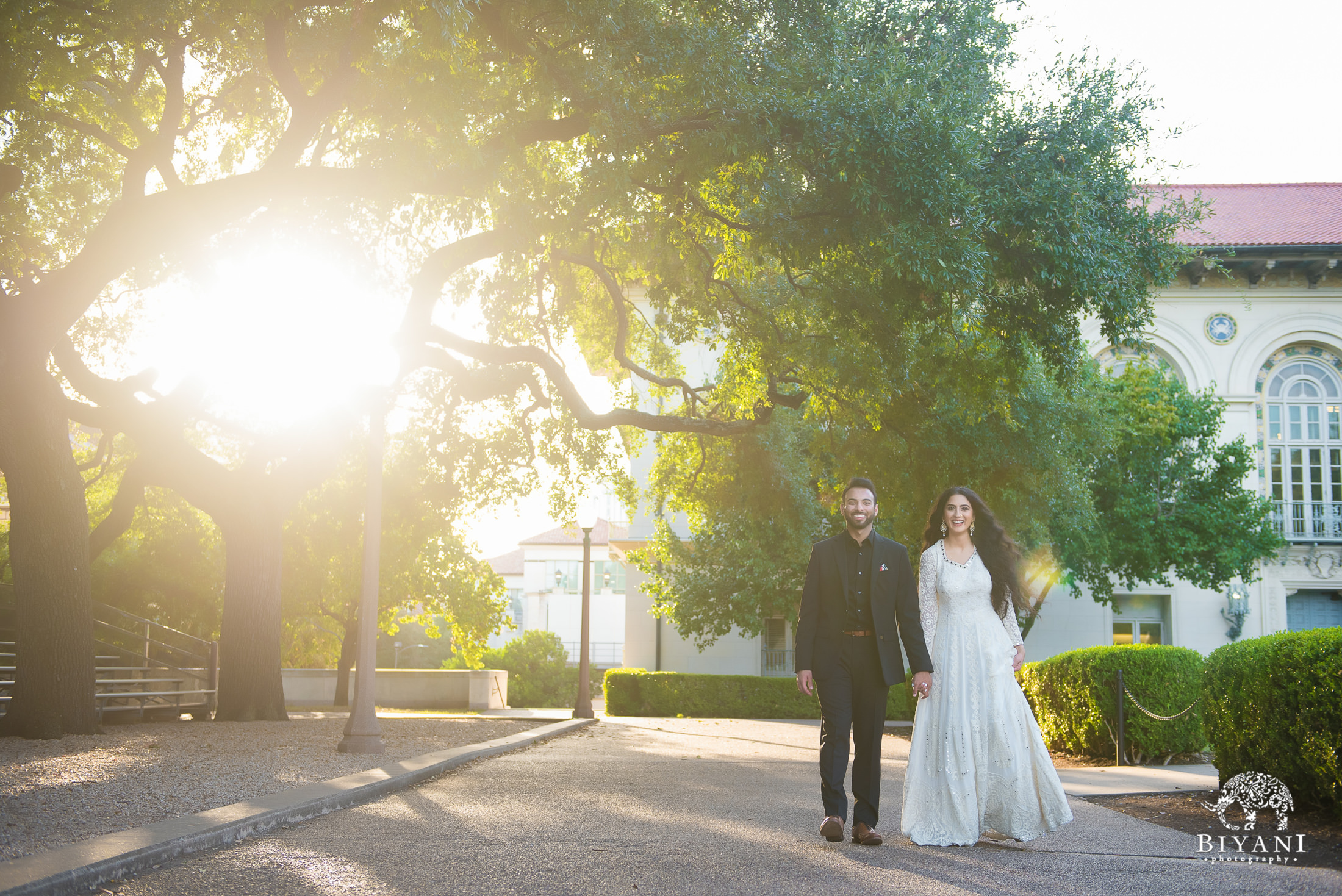 couple walking on sidewalk with sun shining through trees behind them on the University of Texas, Austin campus